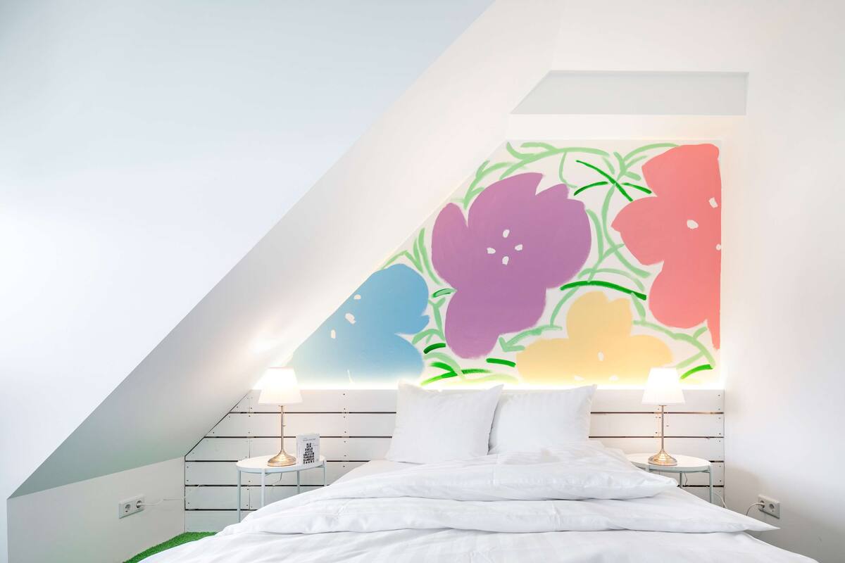 Ein modernes Schlafzimmer mit schrägen Wänden, einem Bett mit weißer Bettwäsche, zwei Nachttischen mit Lampen und einem farbenfrohen Blumenbild an der Wand.