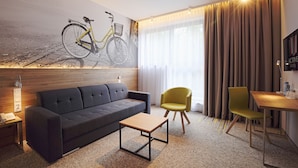 Una habitación de hotel moderna con un sofá, una mesita, una silla amarilla, un escritorio de madera y un mural con motivos de bicicleta en la pared.