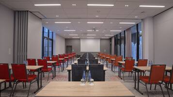 Une salle de conférence avec des rangées de tables et de chaises, de grandes fenêtres et un écran de projection.