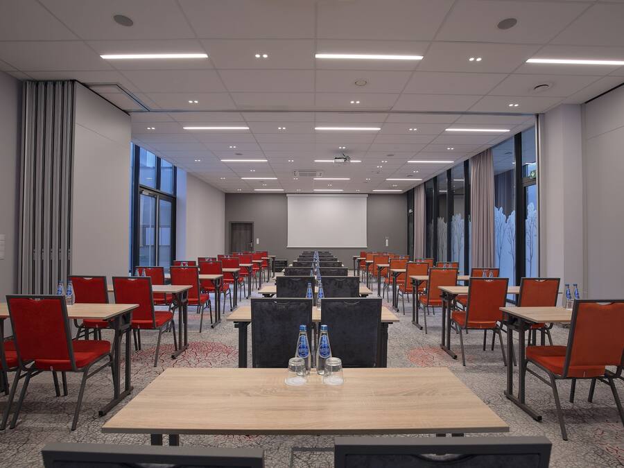 Une salle de conférence avec des rangées de tables et de chaises, de grandes fenêtres et un écran de projection.