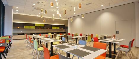 A modern dining area with multiple tables and chairs, overhead lighting, and a kitchen area in the background.