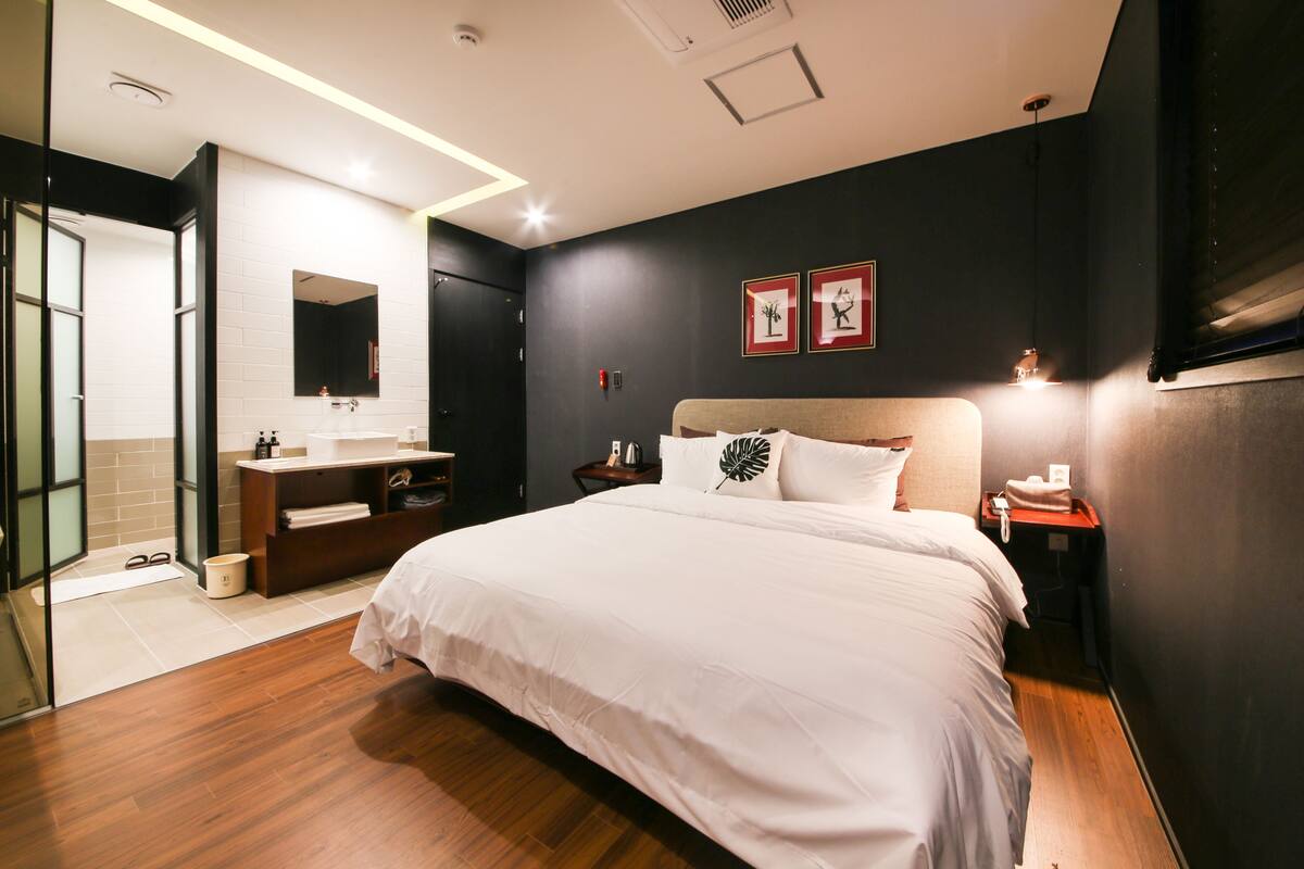 A modern hotel room with a large bed, a bedside table, a nightstand, a mirror, and a small desk with a chair.