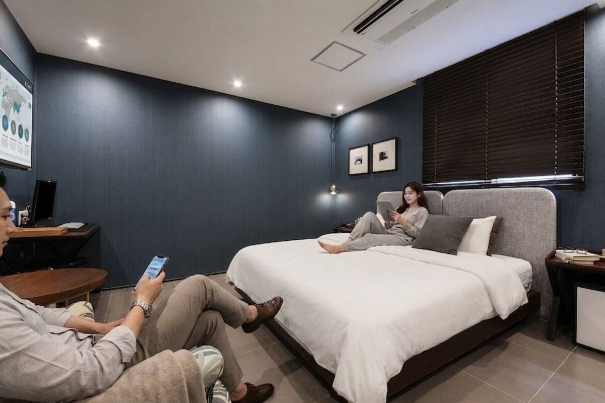 A modern hotel room with a large bed, a desk, and a computer. There is a projection screen on the wall and a window with blinds.