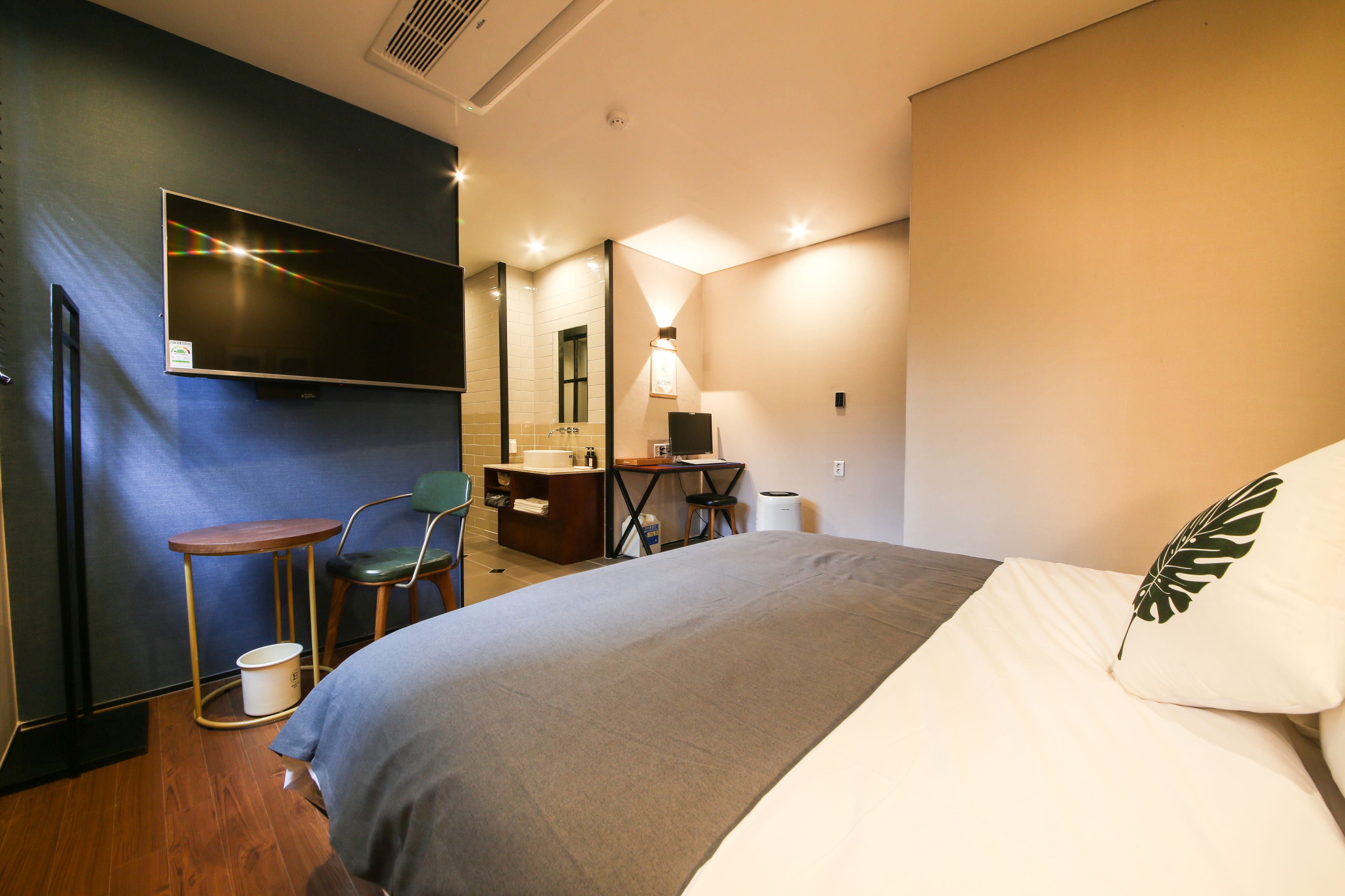 A hotel room with a bed, a desk, a chair, a small table, a bucket, and a TV mounted on the wall.
