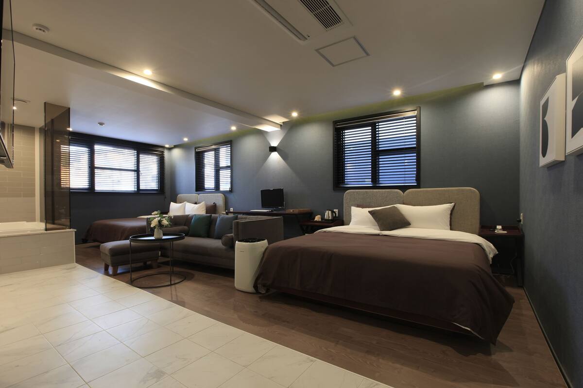 A modern hotel room with a large bed, a sofa, a small table, and a window with blinds.