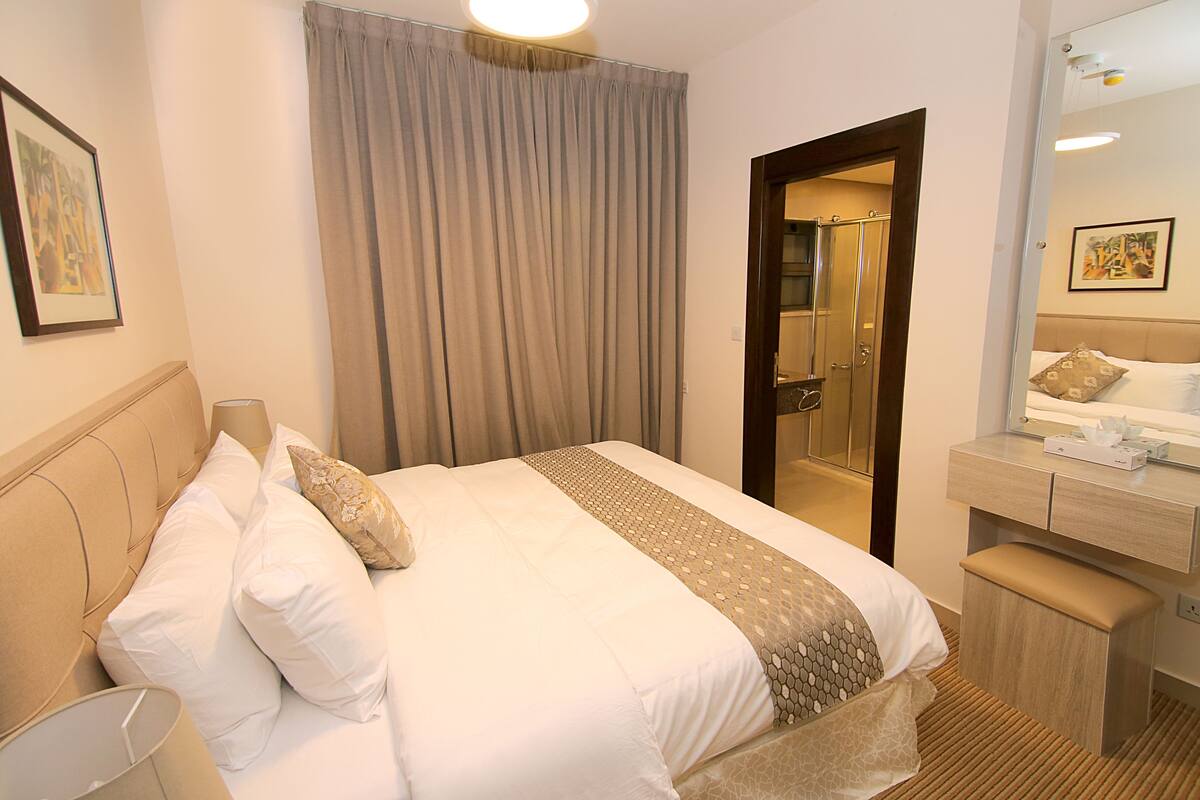 A hotel room with a bed, a bedside table with a lamp, a bathroom visible through an open door, and a mirror above the table.