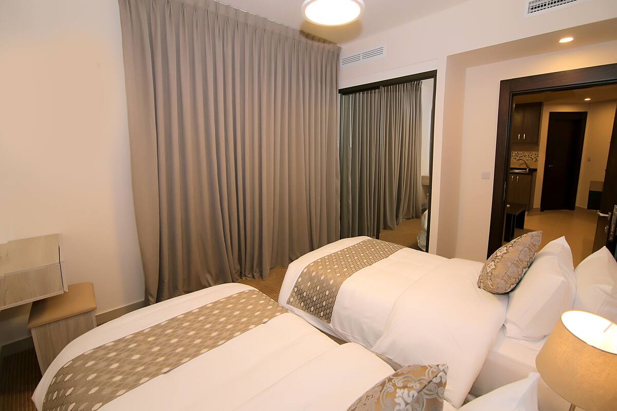 A hotel room with two beds, a bedside table, a lamp, and a view of the hallway.