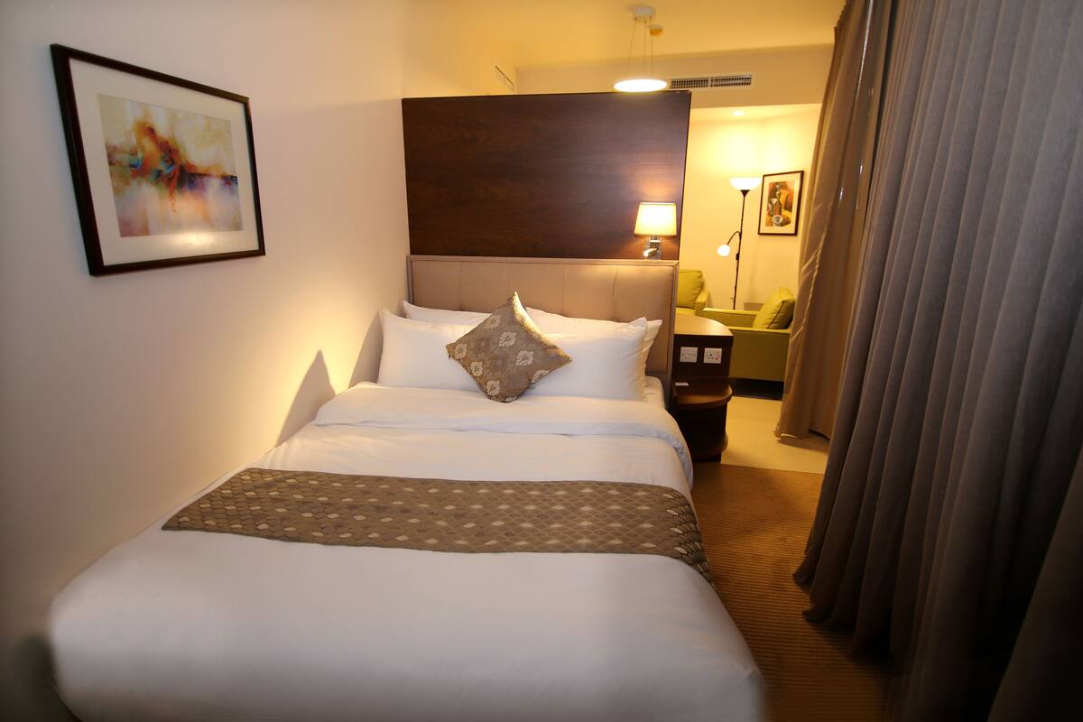 A hotel room with a neatly made bed, a headboard, a bedside table with a lamp, and a view of the hallway.