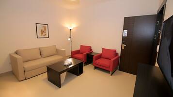 Ein Wohnzimmer mit einer Couch, zwei roten Sesseln, einem Couchtisch, einer Stehlampe und einem Fernseher.