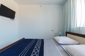 Quarto de hotel simples com uma cama, uma televisão fixada na parede e uma janela com cortinas.