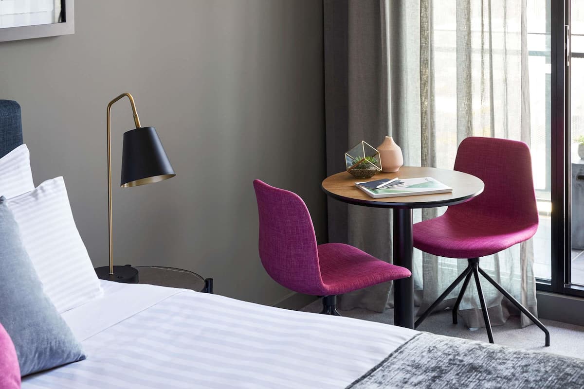 Een hotelkamer met een bed, een bureau met twee stoelen, een lamp en een raam met gordijnen.