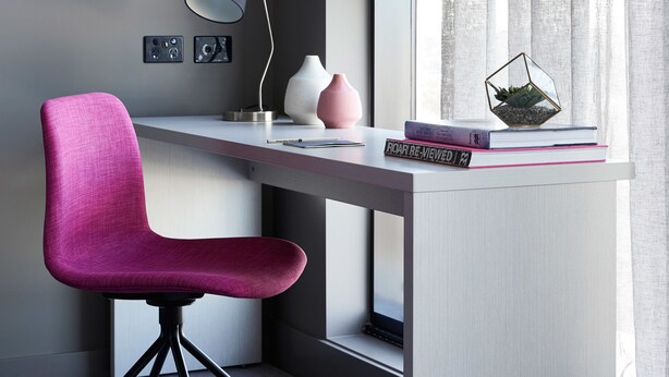 Escritorio moderno con una silla rosa, una lámpara y objetos decorativos, incluyendo libros y un terrario de vidrio.