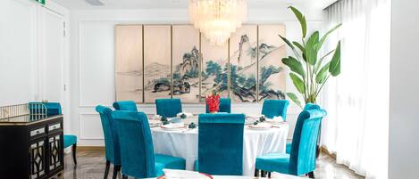 Un comedor con una mesa redonda dispuesta para seis personas, sillas tapizadas en azul, una araña y un cuadro grande en la pared.