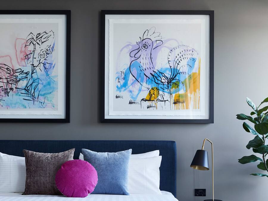 Un dormitorio con una cama, almohadas, una lámpara y dos obras de arte enmarcadas en la pared.
