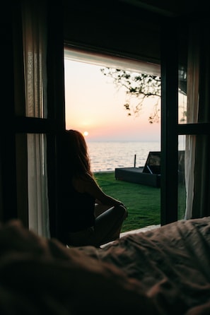 A room with a large window overlooking a grassy area and a body of water at sunset. - Regenta Arie Lagoon Negombo (Negombo)