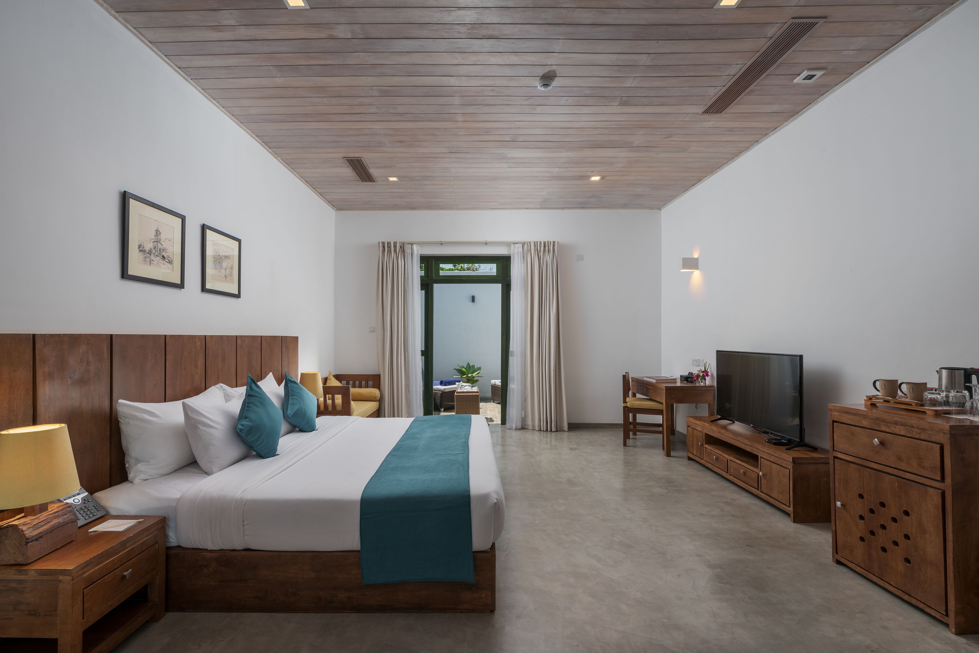 Habitación de hotel con cabecera de madera, cama con ropa de cama blanca y turquesa, mesita de noche con lámpara, televisor sobre un mueble de madera, y vistas al exterior a través de una puerta.
