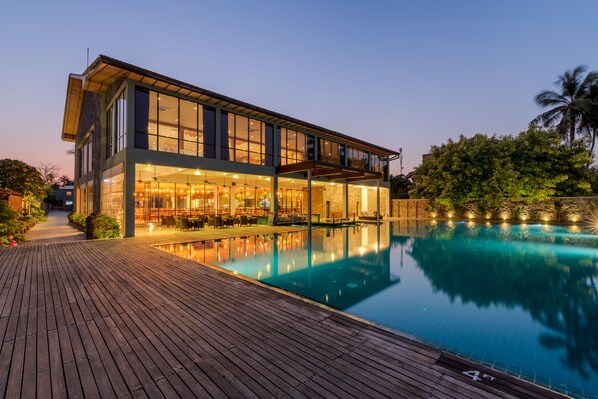 A modern two-story building with a large glass facade, a swimming pool, and a wooden deck. - Regenta Arie Lagoon Negombo (Negombo)