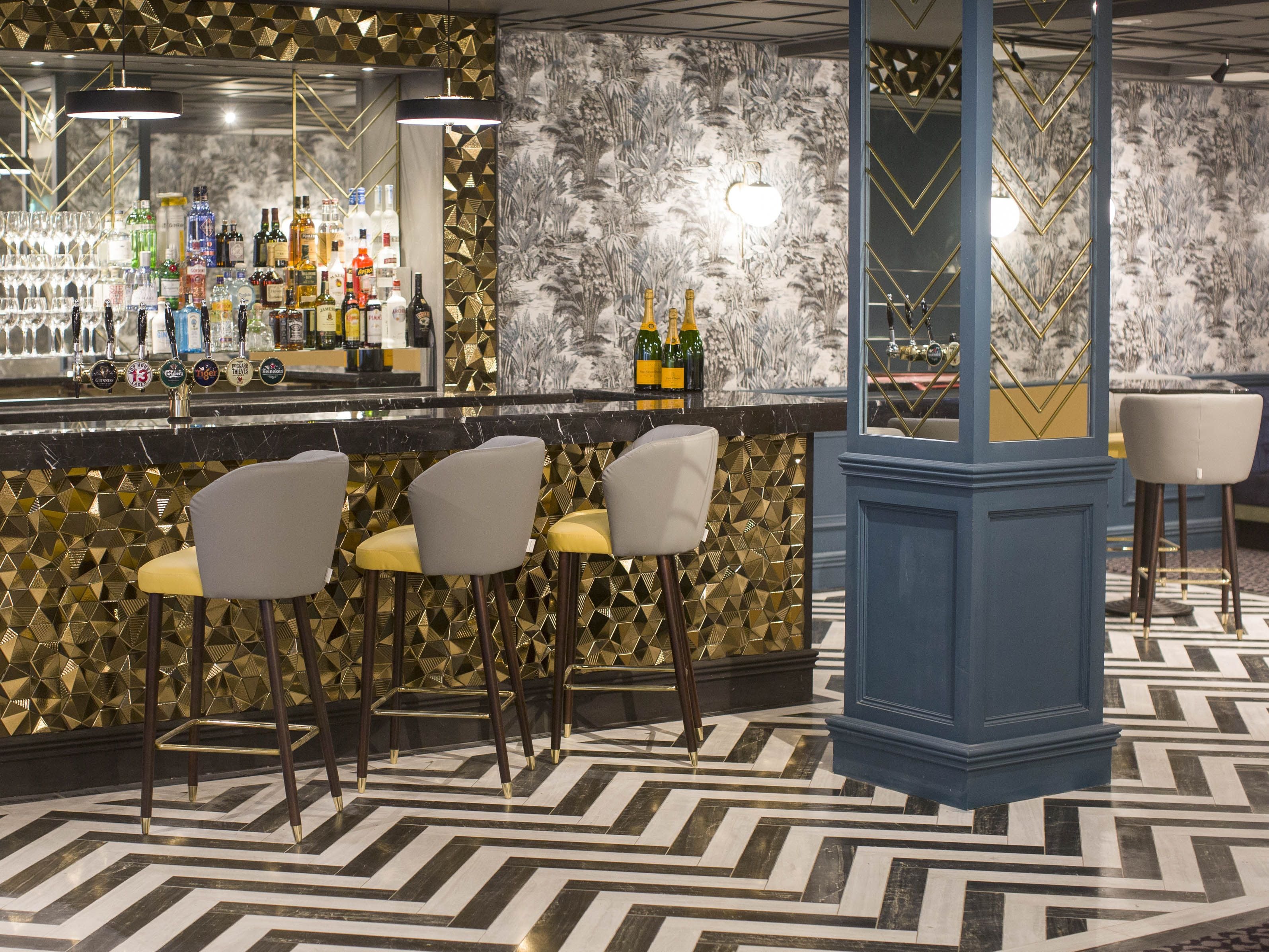 A bar area with a patterned floor, bar stools, and a variety of bottles and glasses on the shelves.