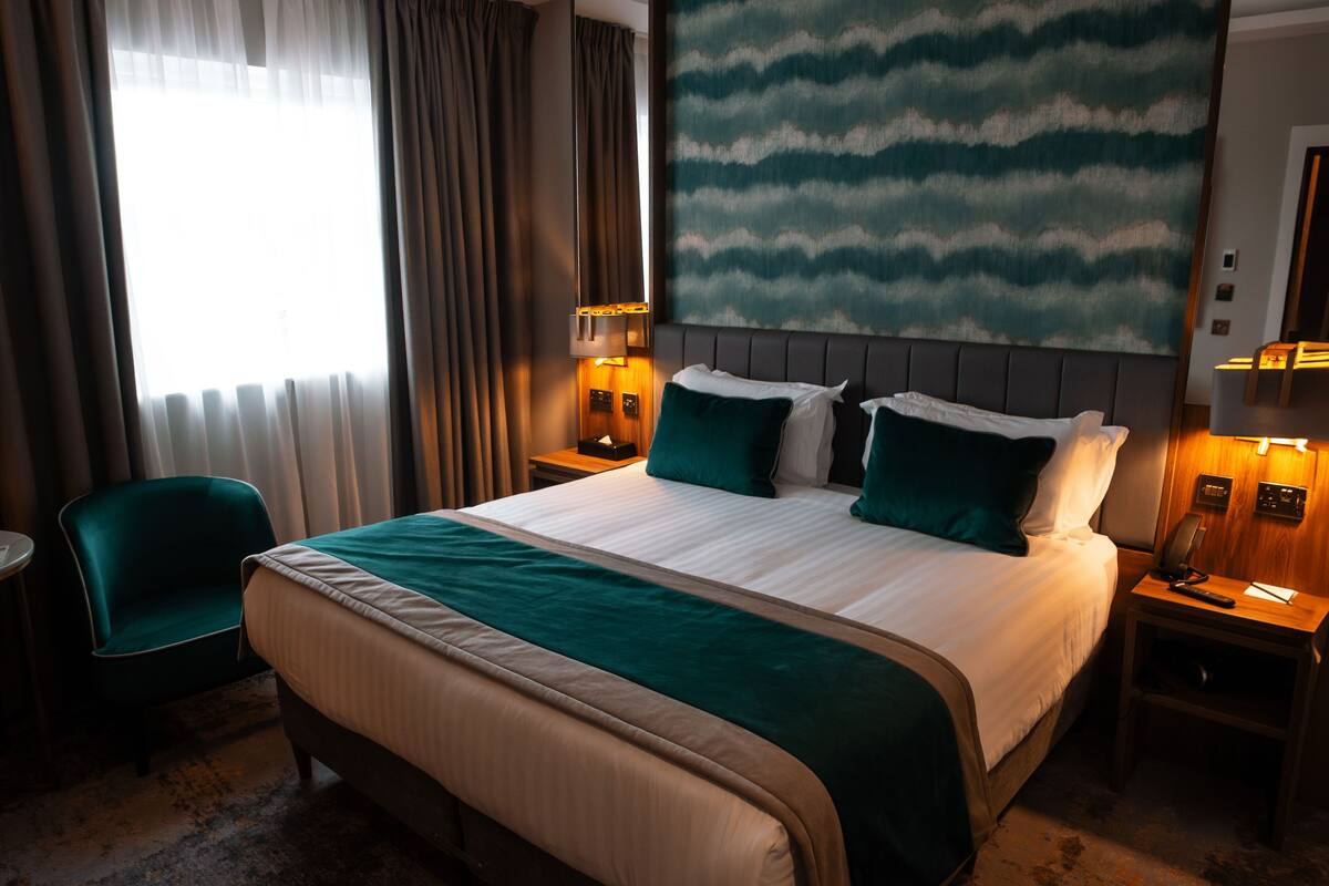 Habitación de hotel con una cama grande, una mesita de noche, una silla y vistas al exterior por la ventana.