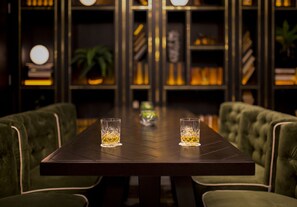 Una mesa con dos vasos de líquido ámbar, junto a sillas tapizadas de verde, en una habitación poco iluminada con estanterías al fondo.