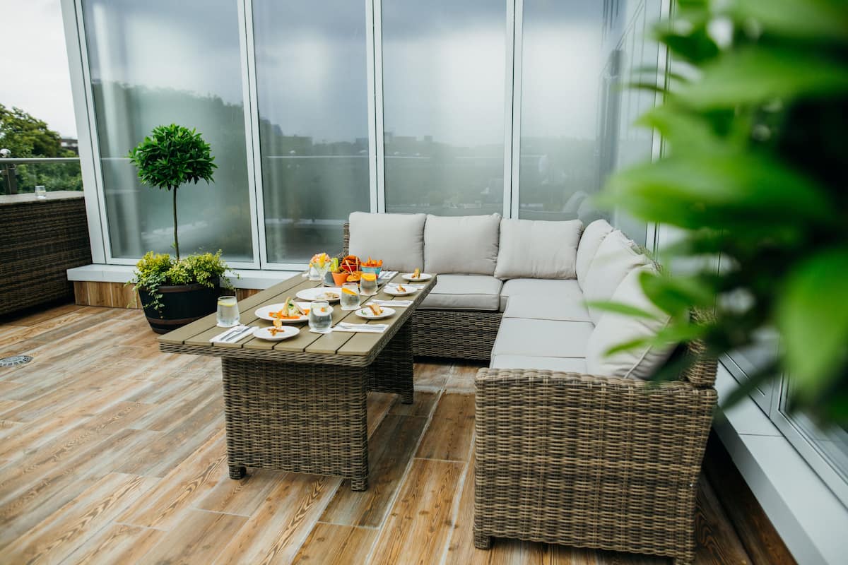 Un área de estar al aire libre, moderna, con una mesa de mimbre dispuesta para comer, un sofá con cojines y una planta en maceta.