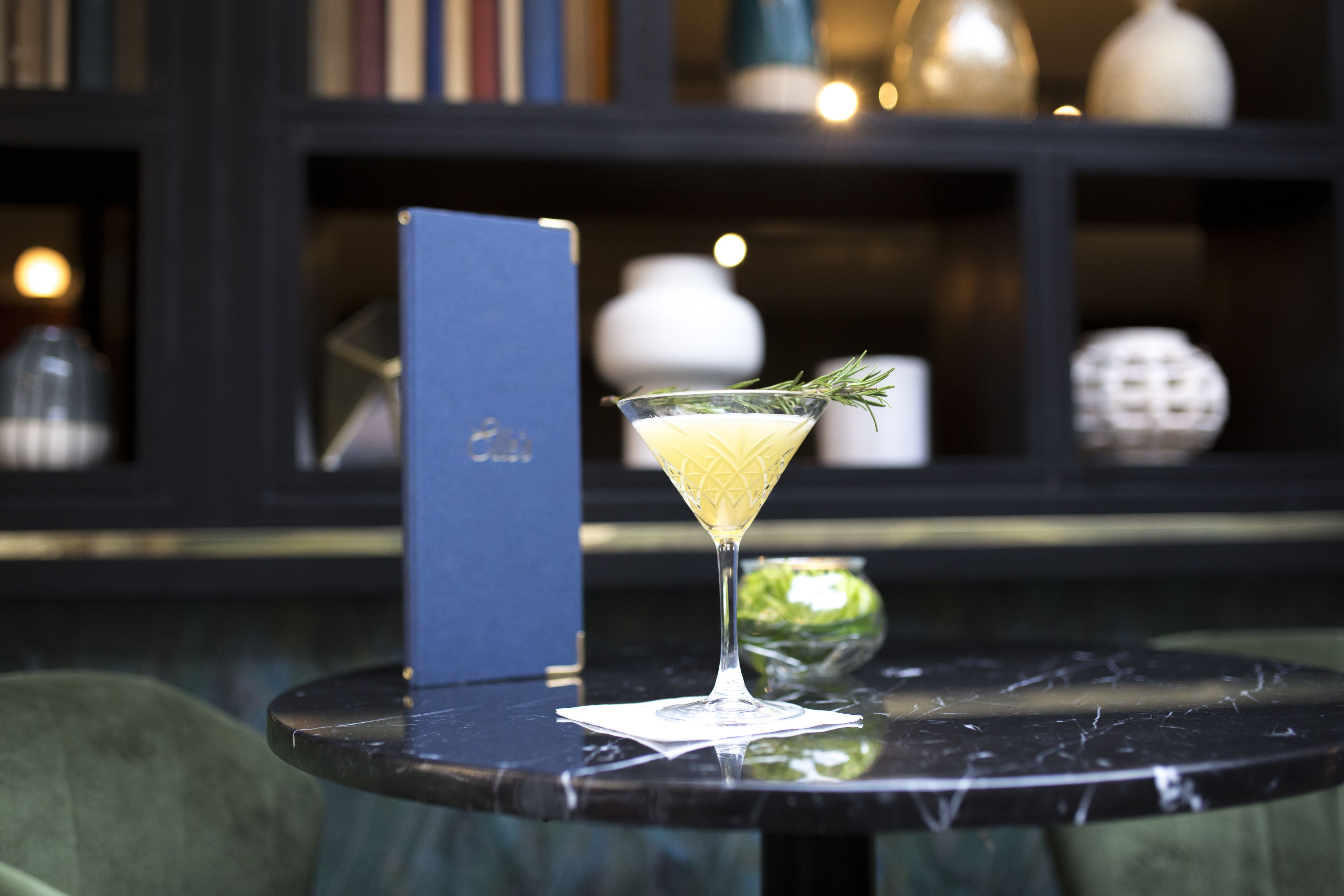 A martini glass with a lemon twist on a marble table, a menu, and a green chair in a dimly lit room with shelves in the background.