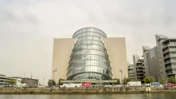 Un edificio moderno con fachada de vidrio curva y techo plano, situado cerca de una masa de agua, con vehículos y otros edificios al fondo.