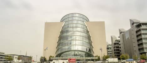 A modern building with a curved glass facade and a flat roof, located near a body of water with vehicles and other buildings in the background.