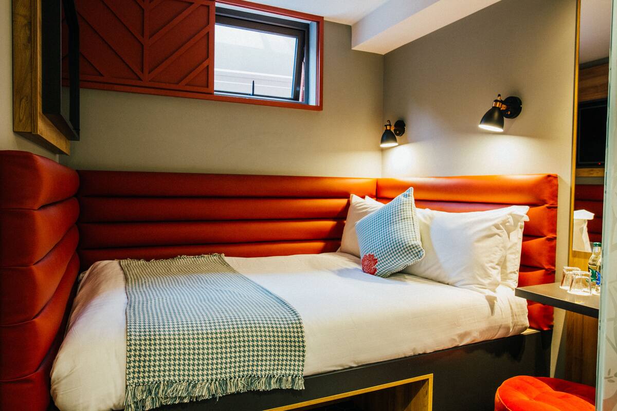 Una cama con cabecera tapizada en rojo, ropa de cama blanca y una manta de cuadros.