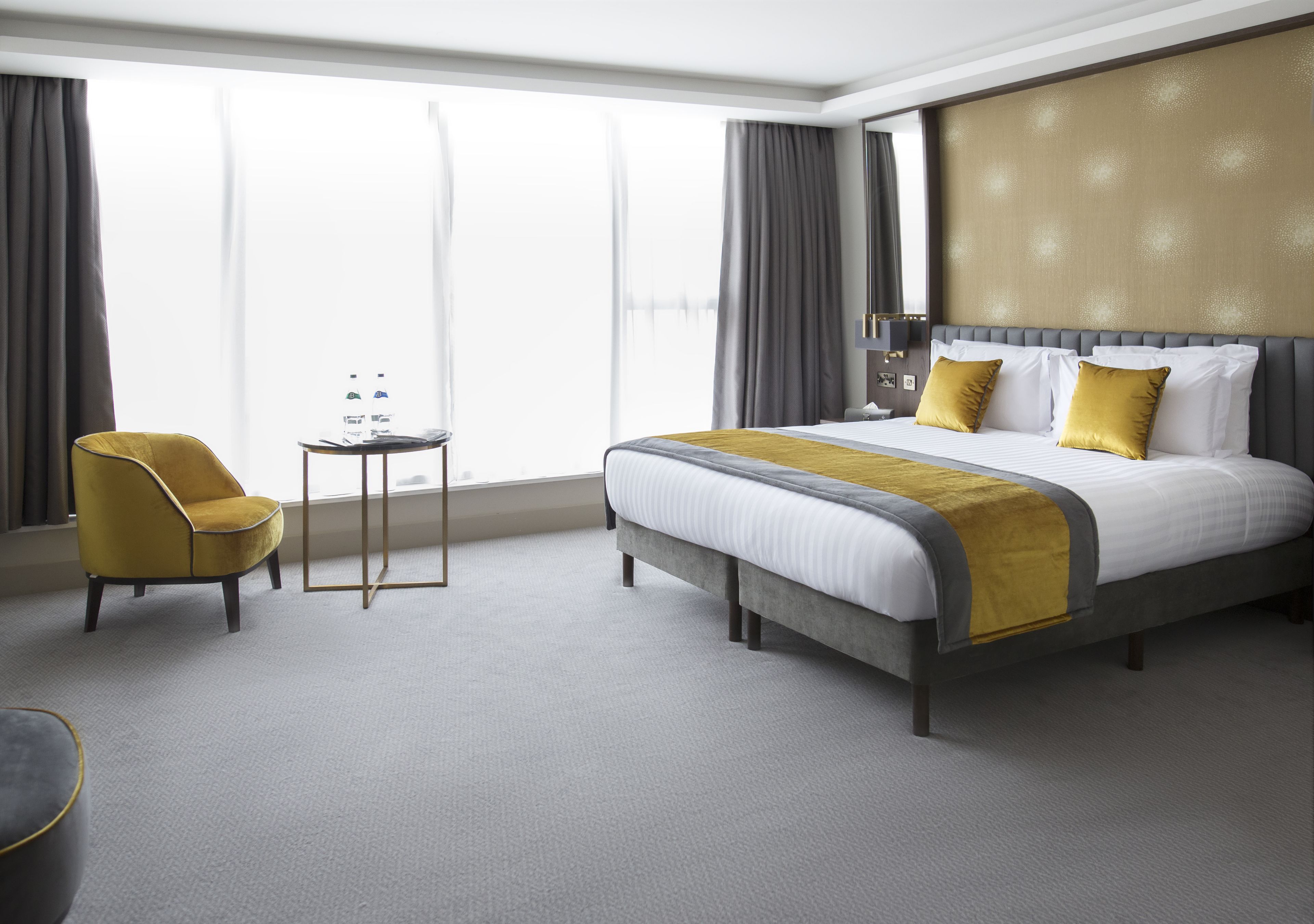 Habitación de hotel con una cama grande, una mesita pequeña, una silla y un ventanal amplio.