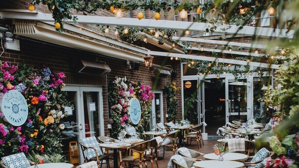 An outdoor cafe with wicker chairs and round tables, surrounded by brick buildings and adorned with hanging lights and flowers. - Iveagh Garden Hotel (Dublin)