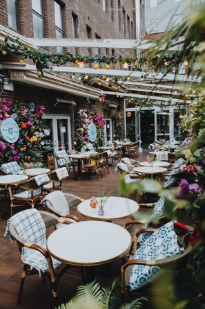 An outdoor cafe with wicker chairs and round tables, surrounded by brick buildings and adorned with hanging lights and flowers. - Iveagh Garden Hotel (Dublin)