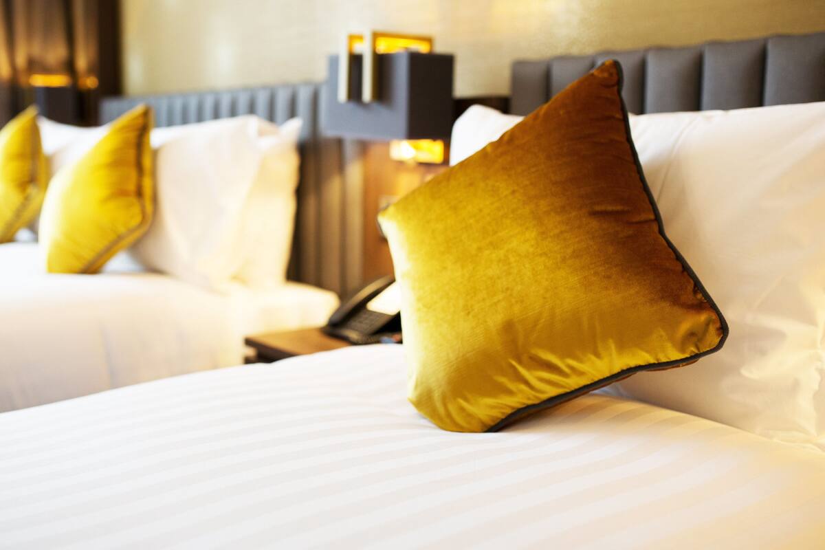Habitación de hotel con dos camas, cada una con ropa blanca y varios almohadones, incluyendo uno dorado destacado.