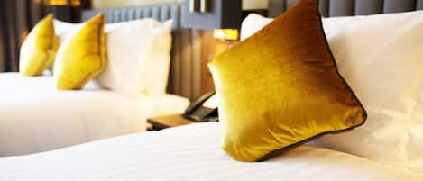 Quarto de hotel com duas camas, cada uma com roupa de cama branca e várias almofadas, incluindo uma almofada dourada em destaque.