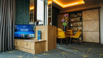 Zona de recepción de un hotel con un mostrador de madera, un televisor mostrando un programa y un área de descanso con sillas amarillas.