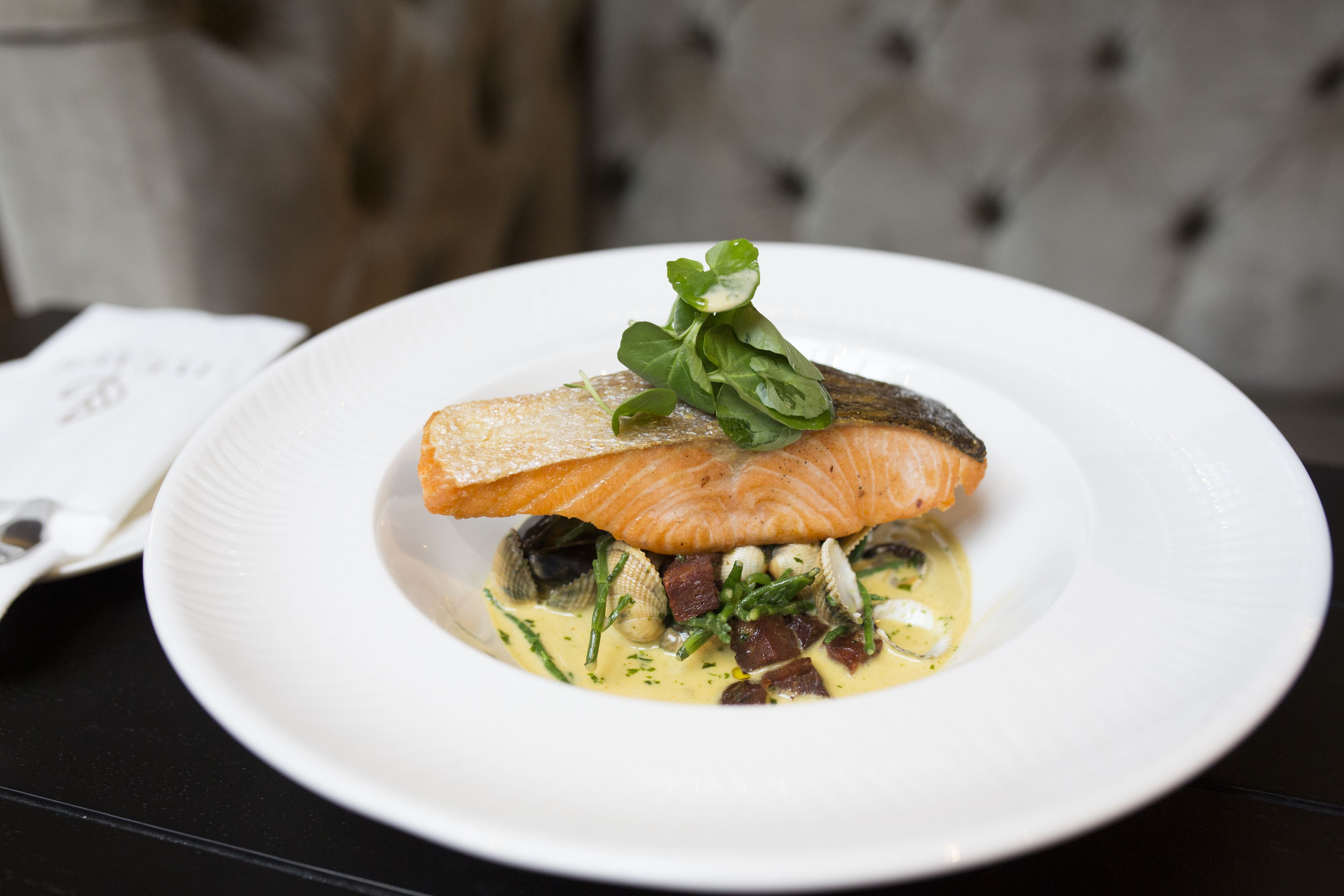 A white plate with a piece of salmon, clams, and greens on a dark table.