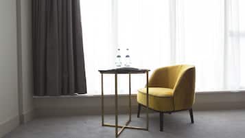 A room with a yellow armchair, a small table with two bottles, and a window with sheer curtains.