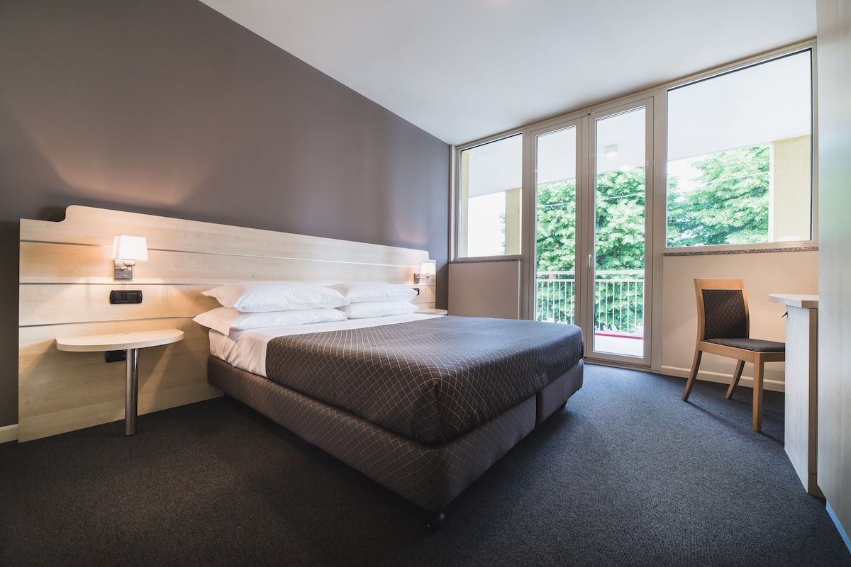 A modern hotel room with a large bed, bedside table, and a desk with a chair.