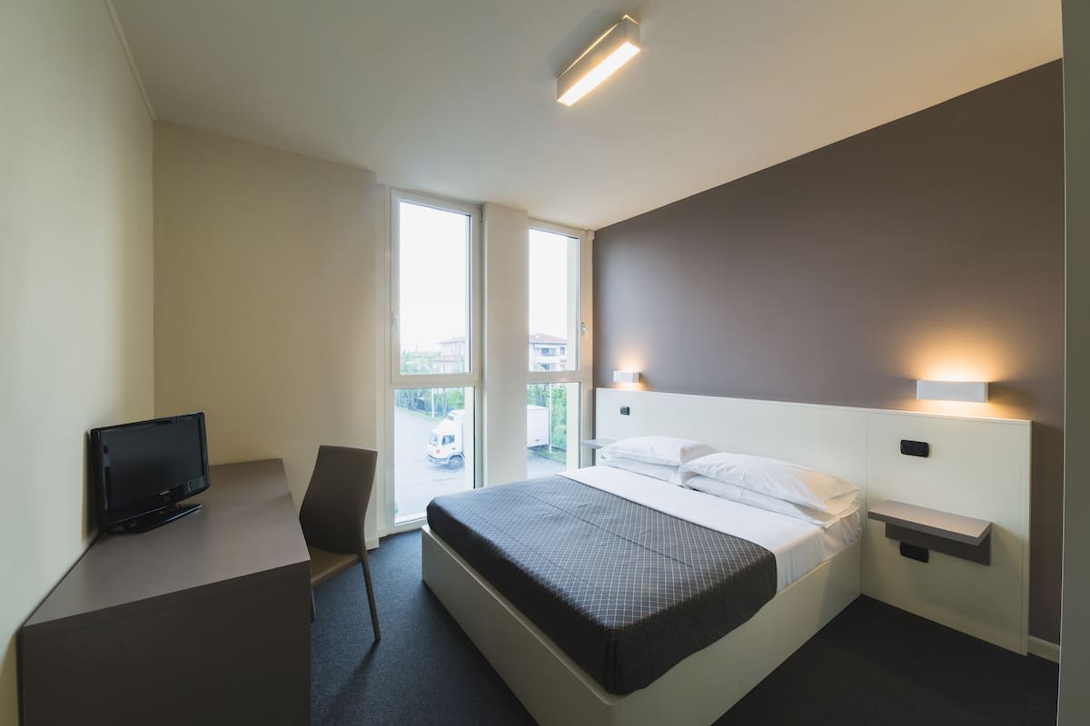 A hotel room with a bed, a desk with a television, a chair, and a window with a view of the outside.