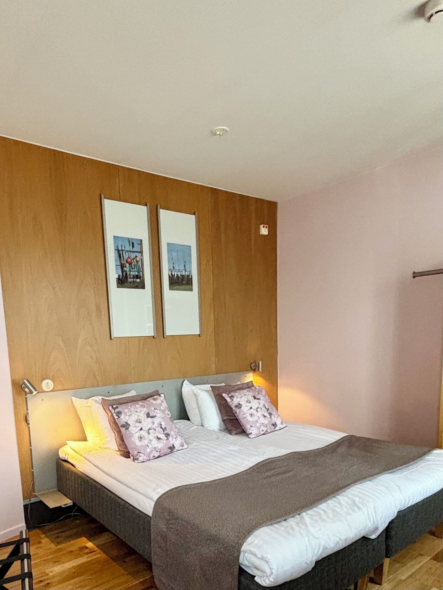 Makuuhuoneessa on puinen sängynpääty, valkoisilla ja ruskeilla tekstiileillä varustettu sänky, kaksi kehystettyä kuvaa seinällä ja yöpöytälamppu.
