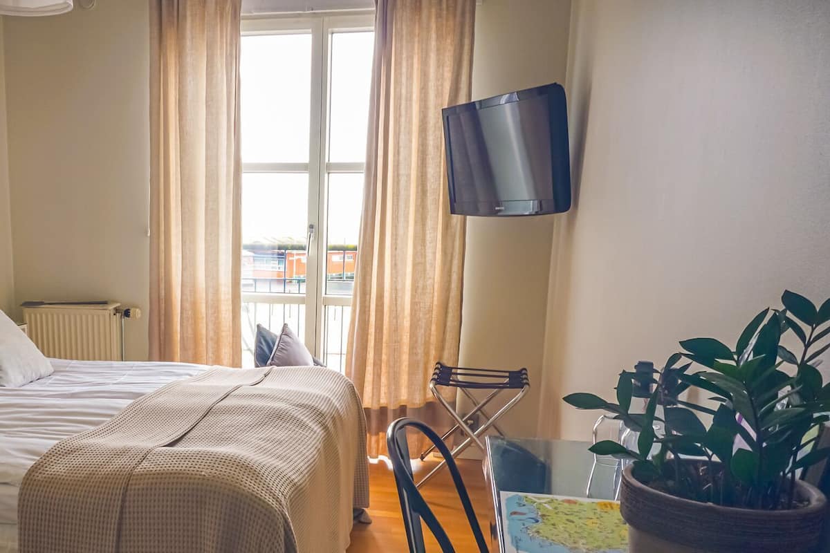 Ein ordentlich eingerichtetes Hotelzimmer mit Bett, Stuhl, Topfpflanze, Flachbildfernseher und einem Fenster mit Vorhängen.