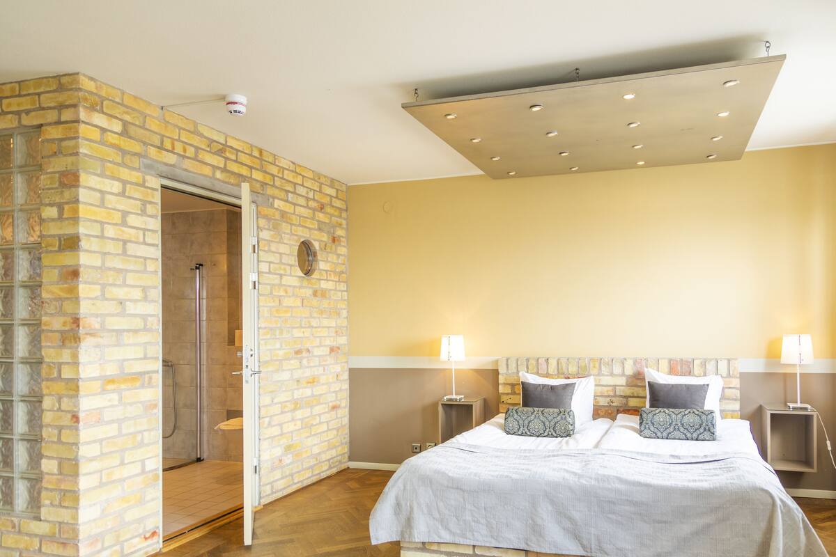 Ein Schlafzimmer mit Backsteinwand, einem Bett mit gemusterten Kissen, Nachttischlampen und einer Deckenleuchte.