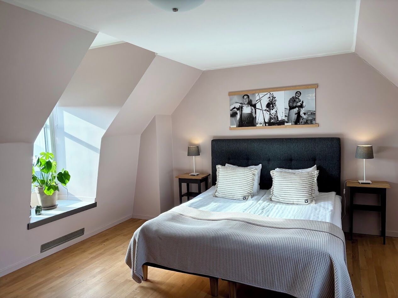 Quarto com teto inclinado, cama com roupa de cama listrada, duas mesas de cabeceira com abajures, janela com planta e um quadro emoldurado sobre a cama.