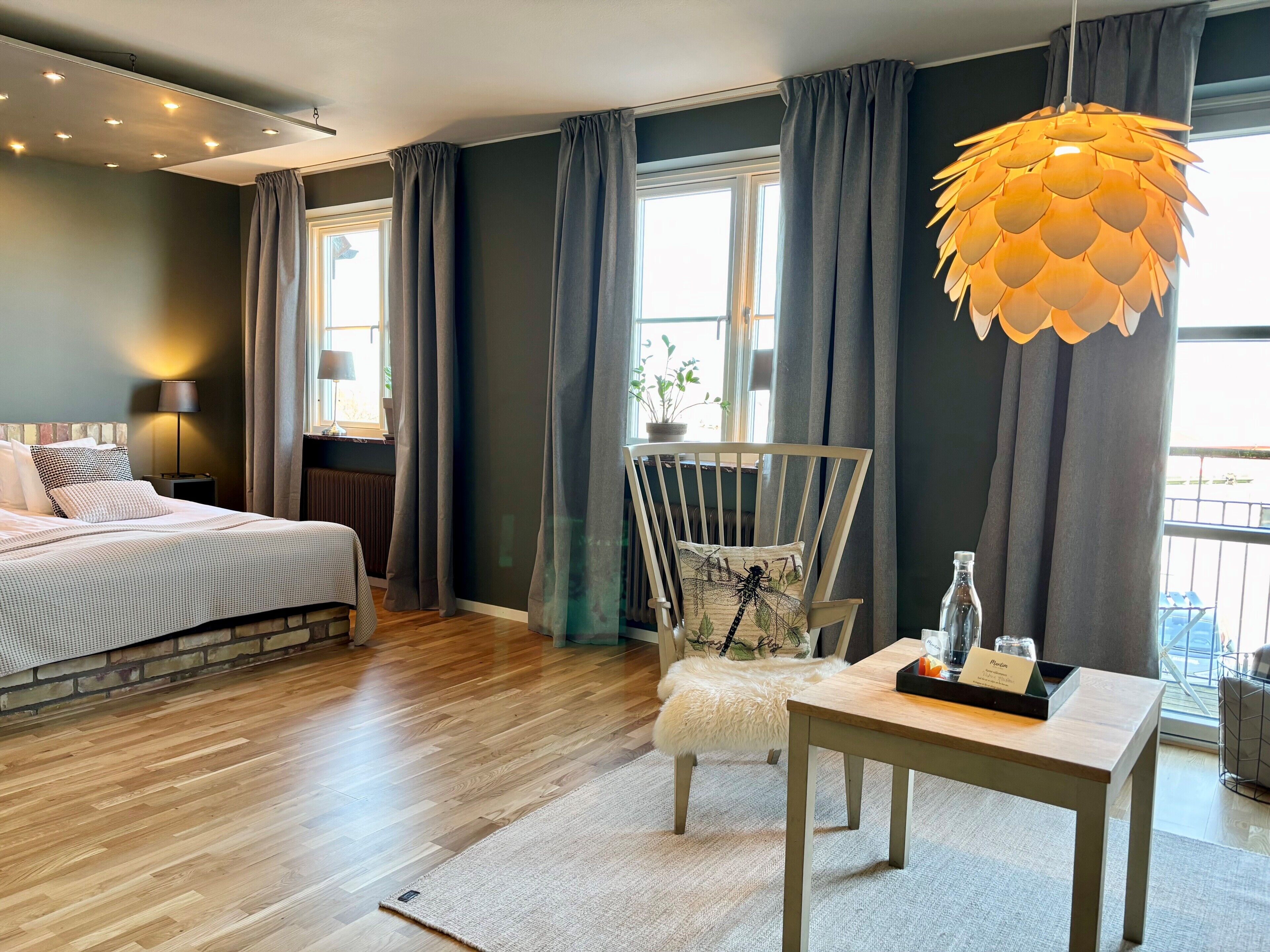 Hotelkamer met een bed, een stoel met een sierkussen in de vorm van een libelle, een tafel met een dienblad, en een hanglamp.