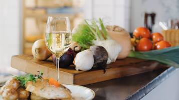 A kitchen counter with a glass of white wine, a plate of food, and various vegetables.