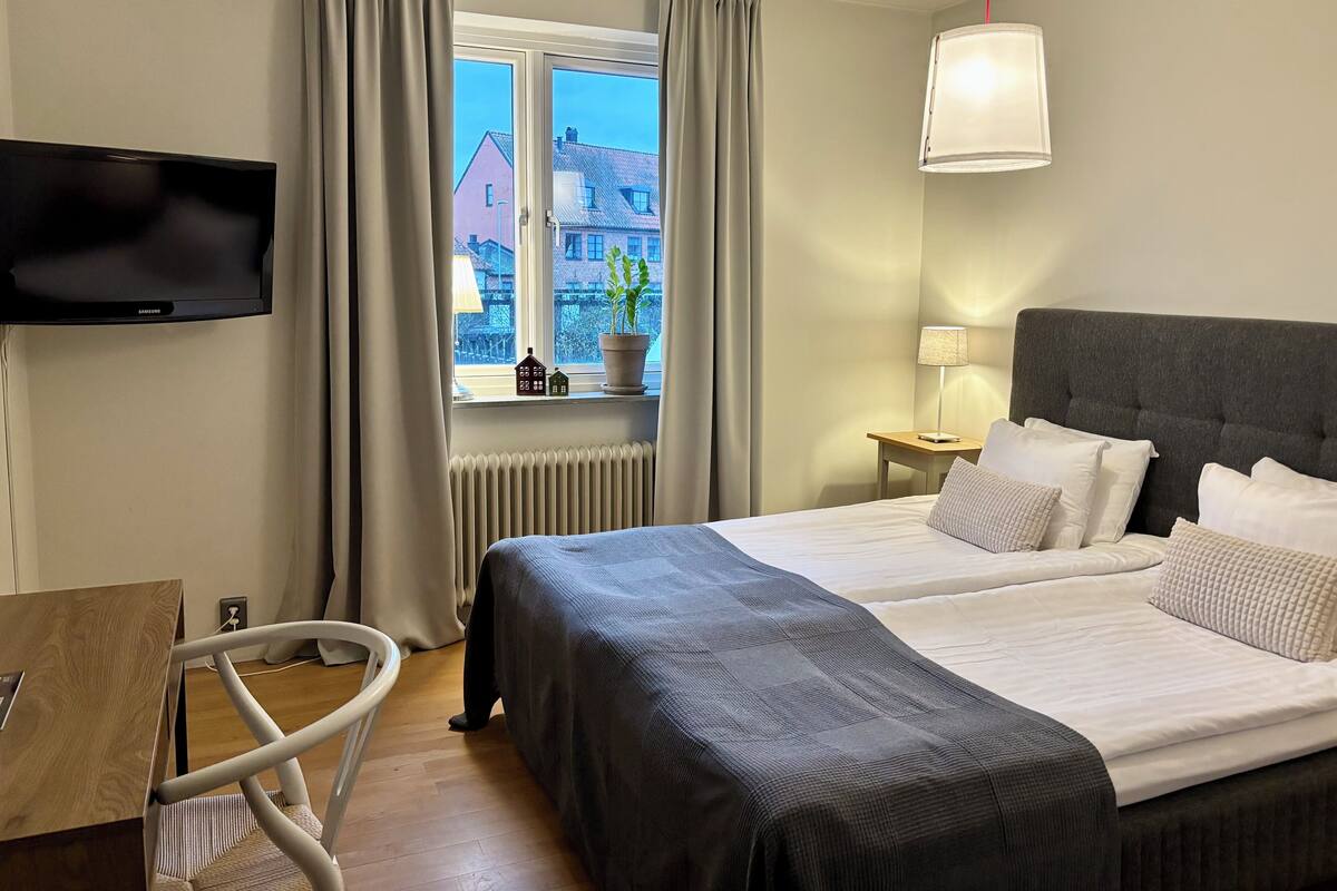 Ein Hotelzimmer mit einem Bett, einem Fernseher, einem Fenster mit Vorhängen, einer Lampe und Blick auf Gebäude.