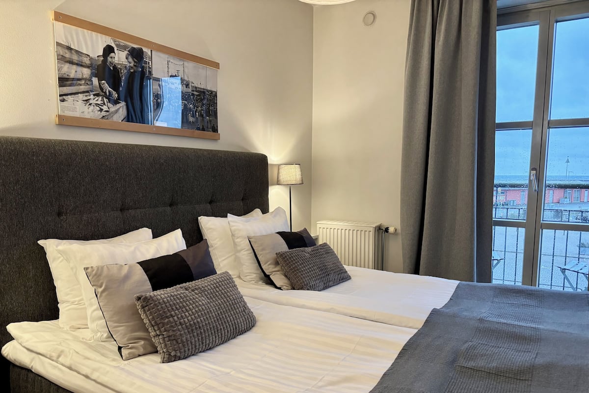 Ein Doppelbett mit weißen Bettwäsche und mehreren Kissen, ein dunkel gepolstertes Kopfstück und ein großes Fenster mit Vorhängen.