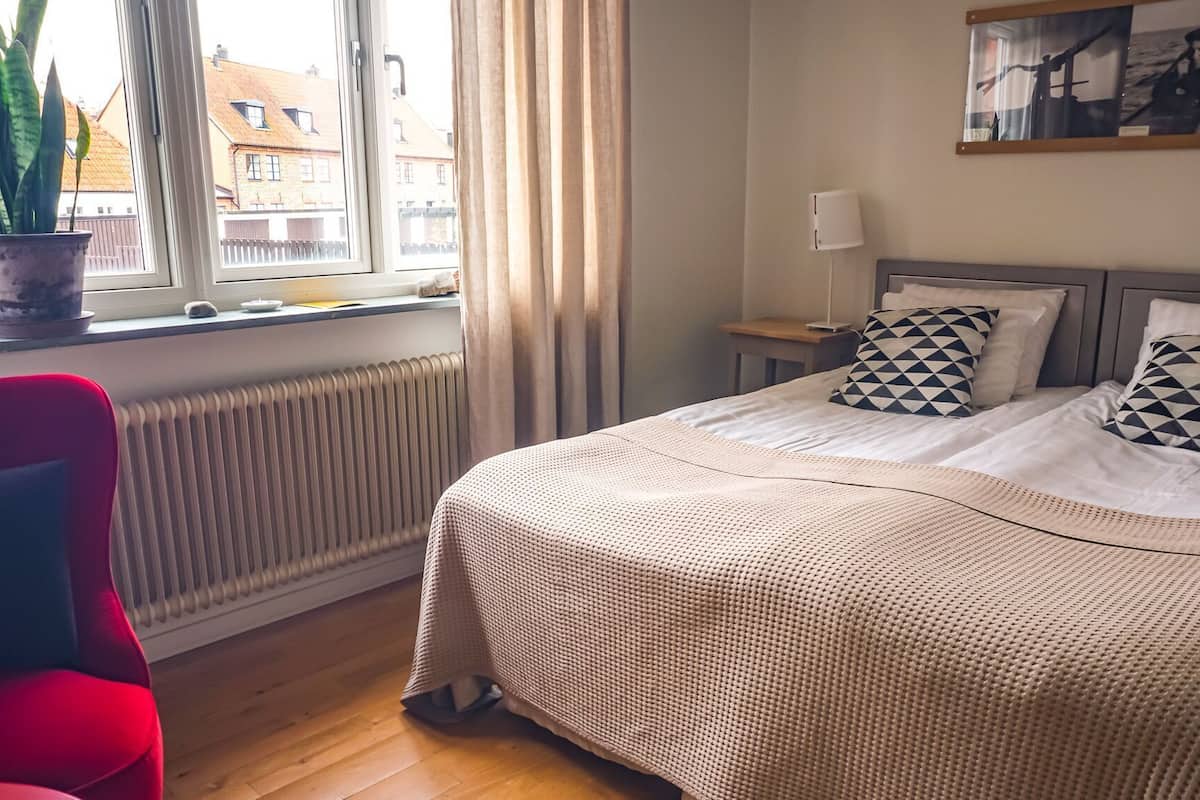 Ein Schlafzimmer mit einem Bett, einem roten Sessel, einer Topfpflanze, einem Heizkörper und einem fenster mit Vorhängen.