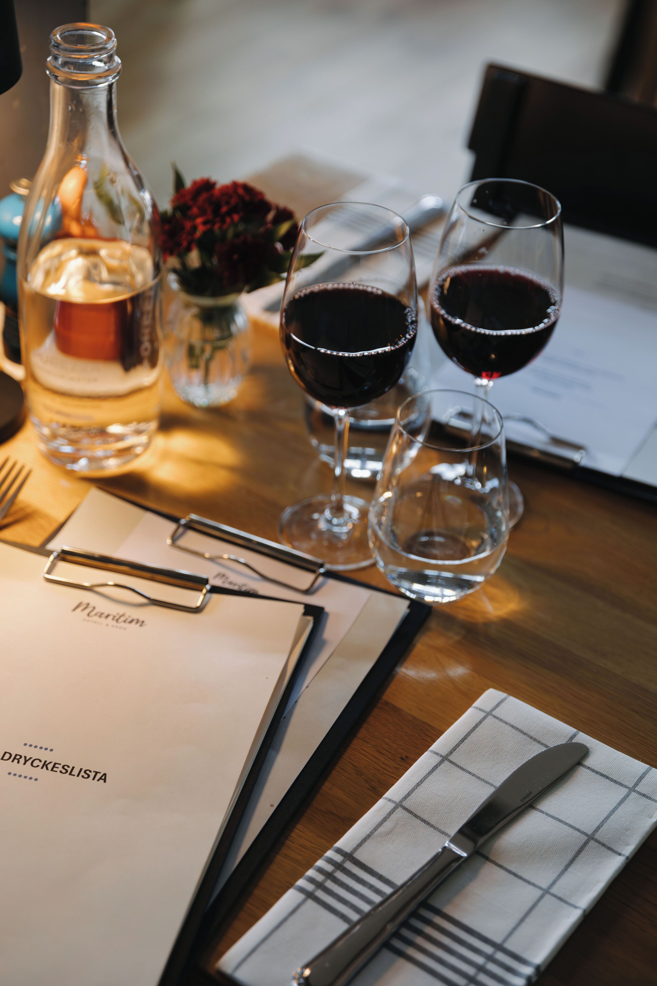 A table with a bottle of water, two glasses of red wine, a menu, and cutlery.