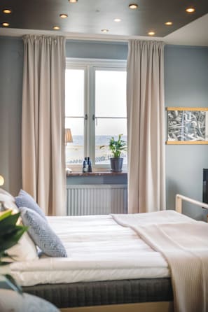 A bedroom with a bed, a window with curtains, a view of the beach, and a potted plant on the windowsill. - Maritim Hotell och Krog (Simrishamn)